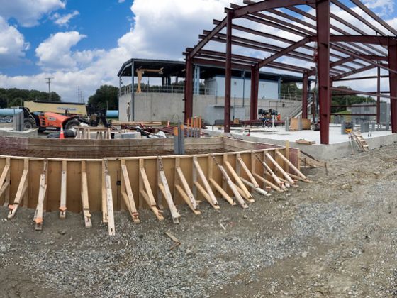 A wooded framework for a building and a metal frame on a concrete platform are shown on a clear day outdoors.