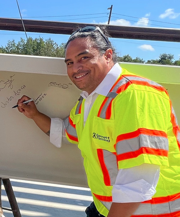 A man in a safety vest writing his name on a white steel beam, smiling for the camera.