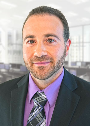 A man with dark hair in a black suit with a purple shirt and black and white plaid tie smiling for the camera.