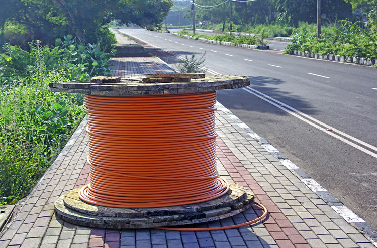 A spool of orange fiber optic broadband cable sitting on a brick sidewalk along a roadway.