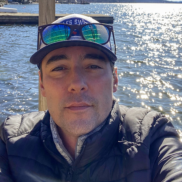 Bryan is sitting on a dock enjoying a beautiful, sunny day on the lake. He is wearing a hat, sunglasses on his head, and a black puffy jacket.