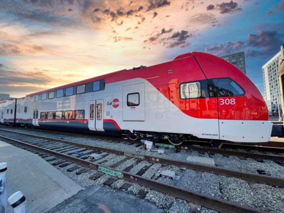 A white electric train with bright red accents sits still on the tracks with hazy skies overhead.