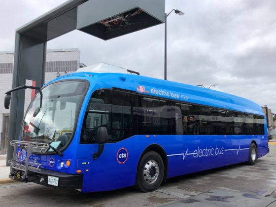 A blue electric bus parked beneath an overhead charge port.