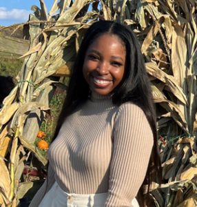 Construction Services Project Manager Emani Majors stands outside smiling in front of some corn stalks on a sunny day while wearing white shorts and a cream-colored long-sleeve shirt.