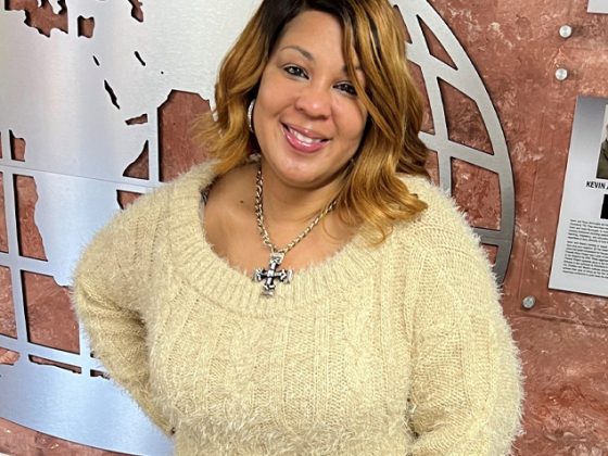 Cybersecurity expert Tamika Bass smiles at the camera with her right hand on her hip while wearing a tan sweater in front of a brown wall.