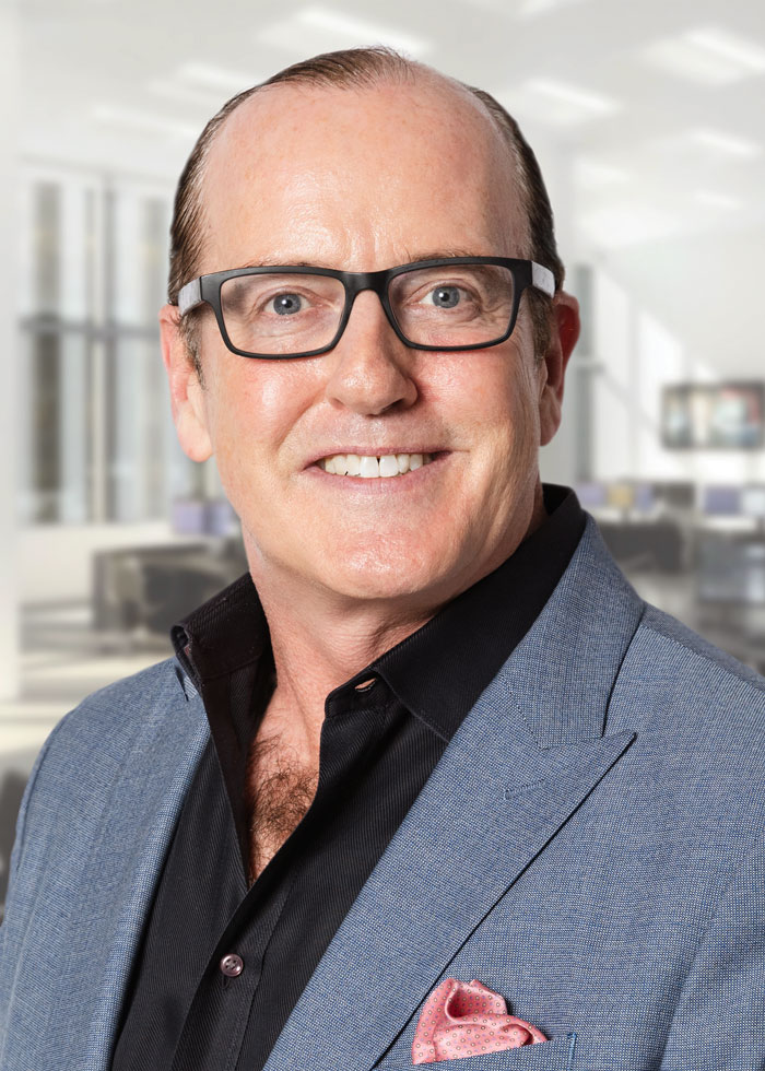 A man with black glasses wearing a gray suit and black shirt smiling for the camera.