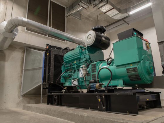 A large green generator is centered on a concrete platform in a room with silver ducting venting it to the exterior.