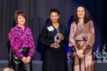 Three women stand smiling on a stage with a black curtain behind them. The woman in the center is holding a glass award.