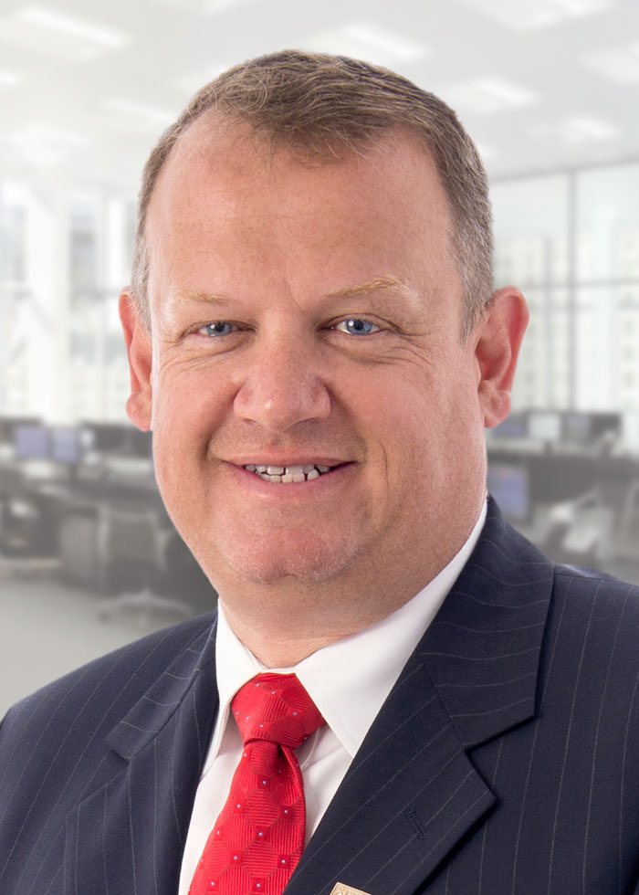 A portrait of Andy Gillespie smiling. He is wearing a dark, pinstriped suit jacket, white button-up shirt, and a red tie.