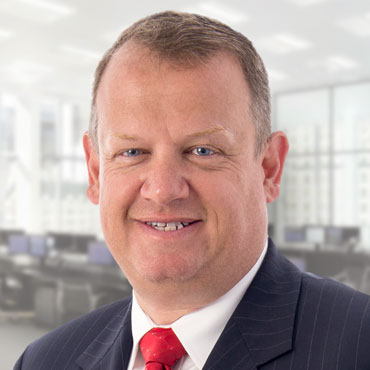 A portrait of Andy Gillespie smiling. He is wearing a dark, pinstriped suit jacket, white button-up shirt, and a red tie.