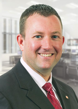 A man with brown hair wearing a black suit jacket, white shirt and red tie smiling for the camera.