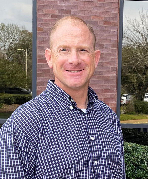 A Caucasian man in blue plaid button-down shirt smiles outdoors for a picture.