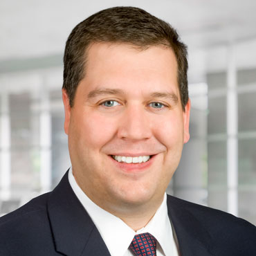 A man with brown hair in a white shirt with a red and blue tie and black suit jacket smiling for the camera.