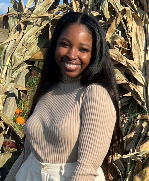 Construction Services Project Manager Emani Majors stands outside smiling in front of some corn stalks on a sunny day while wearing white shorts, and a cream-colored long-sleeve shirt.