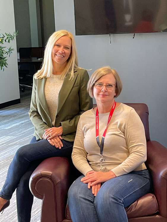 Ruthann Kyler sits in a chair next to her mentor, Heather Eickhoff, who sits on the arm of the chair. They are in an office building smiling.