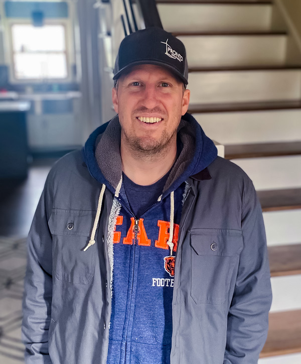 A man wearing a gray ballcap stands in front of a staircase.