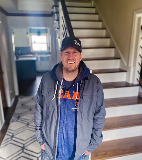 A man wearing a gray ballcap stands in front of a staircase.