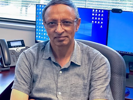 Qasir sits with his arms folded in an office chair in front of two large windows with open blinds. There are two computer monitors on his desk.