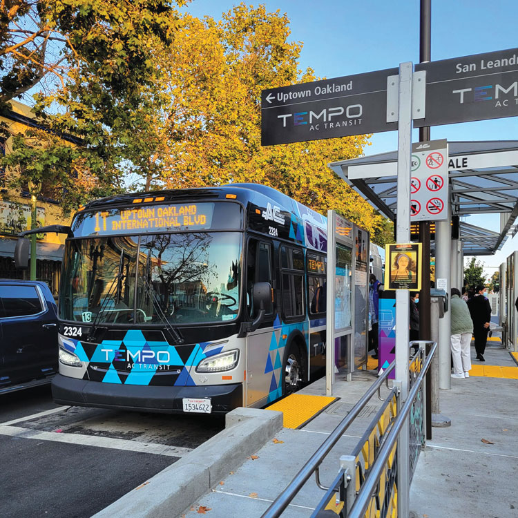 Front view of a blue and silver AC Transit Tempo bus picking up passengers at a BRT station, with wayfinding signage in the foreground.